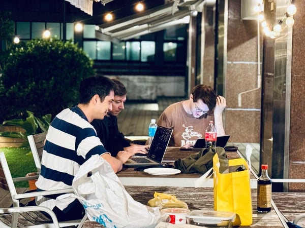 A nighttime photograph captures three men working on laptops and phones around a cluttered wooden outdoor table, illuminated by warm string lights hanging overhead.