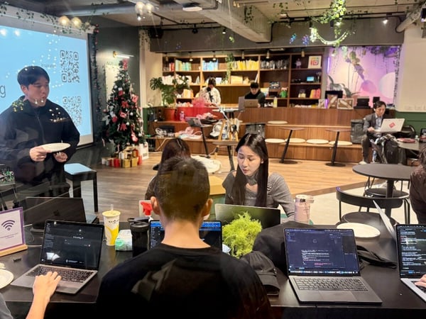 A candid photograph captures a technical meetup or hackathon event taking place indoors, featuring a presenter standing next to a screen displaying 'Hacking' while several attendees work on open laptops at tables. The room is decorated with plants and a Christmas tree, suggesting a relaxed, festive environment.