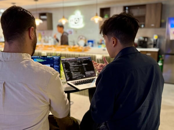 Two men are photographed from behind, seated indoors and collaborating over two open laptops displaying code, likely in a casual office or coworking space setting.
