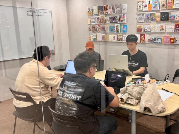 A candid photograph captures three men working on laptops around a table inside a modern office or meeting room, separated by a glass partition visible in the foreground.