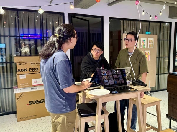 Three men are engaged in a discussion around a high wooden table with open laptops, one displaying a video conference grid, in a casual indoor setting decorated with string lights.