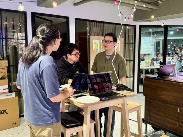 Three individuals are engaged in a discussion around a high wooden table with two open laptops, likely reviewing code or media content in a brightly lit indoor space decorated with string lights.