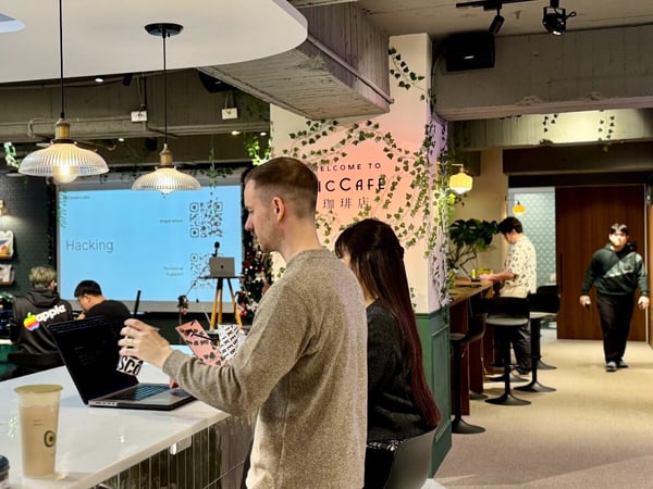 A candid photograph taken inside a modern café or co-working space where a group of people are attending a technical workshop or hacking event, indicated by the word 'Hacking' projected onto a screen in the background.