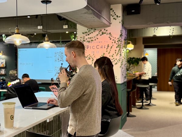 A candid photo captures a man working on a laptop and drinking from a cup at a counter in a modern cafe setting, with a large screen in the background displaying the word "Hacking" and QR codes.