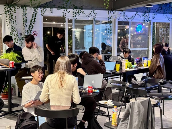 A candid photograph captures a group of young people working intensely on laptops and collaborating around high tables in a dimly lit, casual indoor space, possibly during a hackathon or study session.