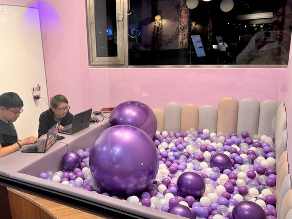 Two individuals are working on laptops inside an unconventional, pink-walled room featuring a large ball pit filled with purple and white balls and several large exercise balls.