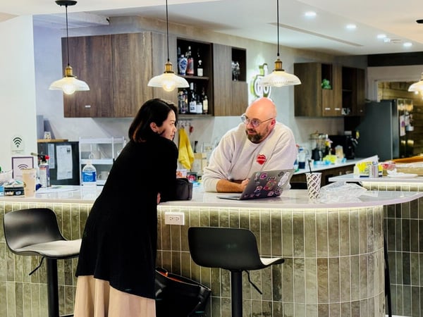 A candid indoor photograph shows a man and a woman engaged in conversation across a curved, tiled bar counter in a modern cafe or breakroom setting, with the man looking at an open laptop.