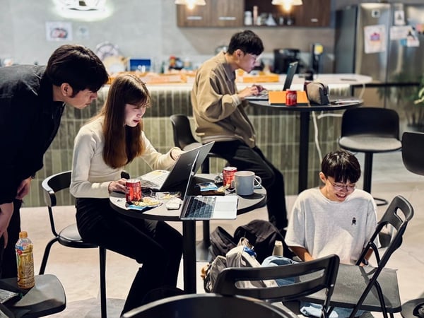 A candid photograph captures four young people working on laptops in a modern, warmly lit indoor setting, likely a co-working space or office kitchen area. A man leans over a woman seated at a small table, collaborating on a screen, while two others work independently nearby.