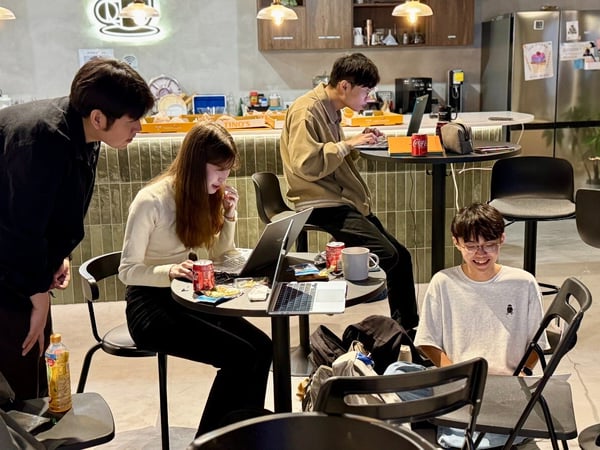 A candid photo showing four young people working on laptops around small, high tables in a casual indoor environment that resembles a cafe or office break room.
