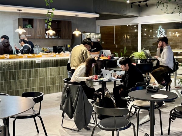 A candid photograph captured inside a modern cafe or co-working space, showing several people seated at small tables working on laptops, with a tiled service counter visible in the background.