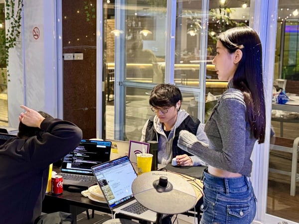 A candid photograph captures a group of four people working around a table in a dimly lit indoor or semi-outdoor setting, characterized by open laptops and warm string lights in the background.