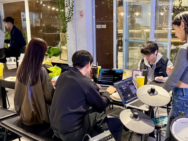 A nighttime photograph captures a group of young adults working on open laptops displaying code at a dark table in a brightly lit indoor/outdoor cafe setting, with parts of a Roland electronic drum kit visible in the foreground.