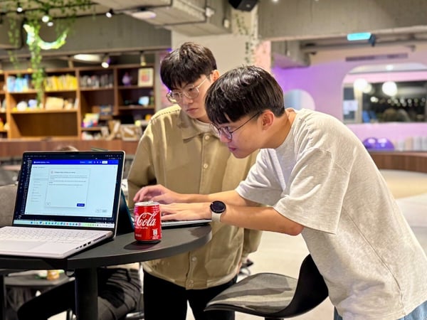 Two young men are leaning over a laptop placed on a small table, actively collaborating or reviewing content in a brightly lit, modern indoor space that resembles a co-working area or library.