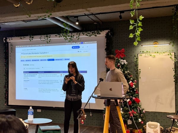A photograph capturing an indoor presentation or demo event, featuring a woman speaking while holding a device in front of a large projection screen displaying the 'ElevenLabs Worldwide Hackathon' website, with a man standing beside her. The venue is decorated with artificial ivy and Christmas decorations, including a decorated tree.