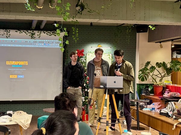 Three young men are giving a presentation or demo in a warmly lit, plant-decorated indoor space, standing in front of a large projection screen displaying the title "ELEVENPOINTS."