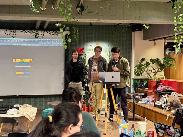 Three young men are giving a presentation or demo of software titled "ELEVENPOINTS GAMES" projected onto a large screen in a casual, indoor event space decorated with plants and holiday accents.