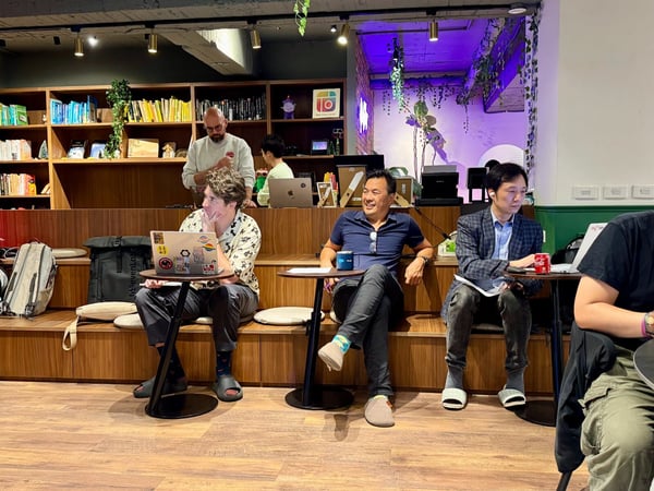 A candid photograph capturing a group of people sitting on tiered wooden seating in a modern, bookshelf-lined indoor space, likely during a casual meeting or workshop.