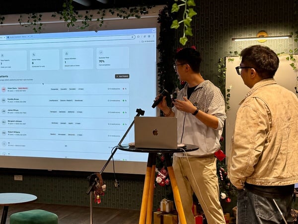 A photo capturing an indoor presentation where a speaker, holding a microphone, gestures toward a large projected screen displaying a patient management software dashboard, while another person stands nearby.