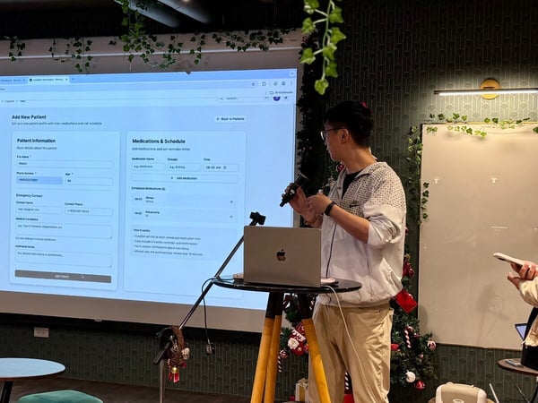 A photo capturing a man giving a software demonstration, projected onto a large screen in a dimly lit, decorated indoor space, showing an 'Add New Patient' form interface.
