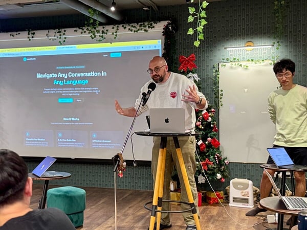 A man is presenting in a casual, decorated indoor space, speaking into a microphone while a large screen behind him displays a website for a language navigation tool called "LevelGuide."