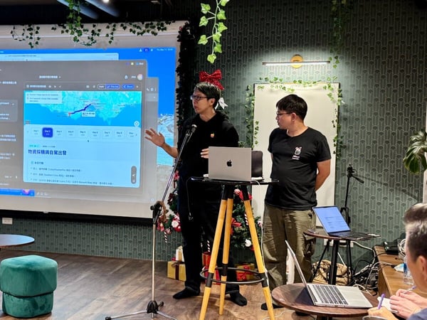 A man is giving a technical presentation indoors, standing next to a large projected screen displaying a web interface with a map and Chinese text, while another man watches nearby.