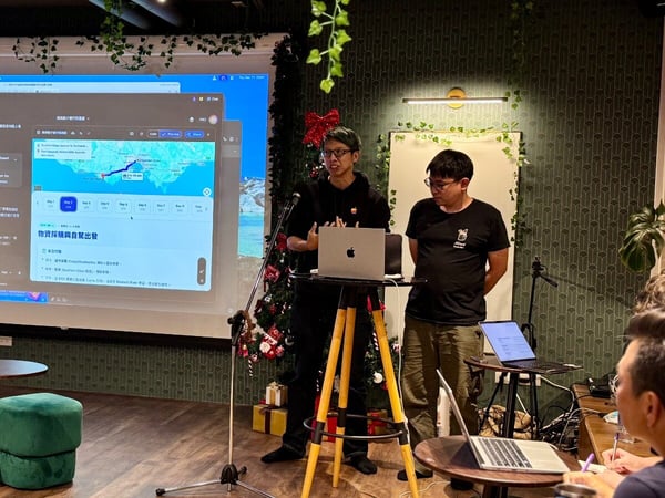 Two men are presenting software or data, featuring maps and text (primarily Chinese) projected onto a large screen in a decorated, indoor venue, while an audience observes from the foreground.