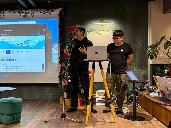 Two men are giving a presentation or technical demonstration indoors, standing near a microphone and laptops, with the content projected onto a large screen behind them.