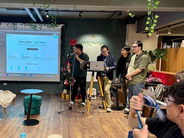 An indoor photograph captures a startup pitch or presentation session, showing several men standing near a projection screen displaying a website interface labeled "Your Links, Your Voice," while an audience member watches from the foreground.