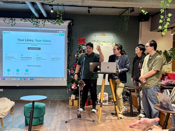 An indoor photo captures a small group of four men attending a presentation, where the speaker on the left gestures toward a projected screen displaying a website interface labeled "LinkHub." The setting appears casual, decorated with hanging plants, patterned wallpaper, and a Christmas tree.