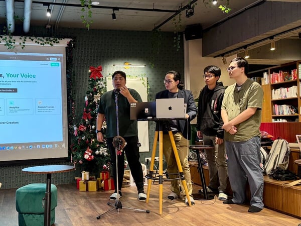 Four young men are gathered indoors, likely giving a product demonstration or pitch, with one man speaking into a microphone while a website interface for "LinkHub" is projected onto a large screen. The indoor setting features warm lighting, a bookshelf, and Christmas decorations including a decorated tree.