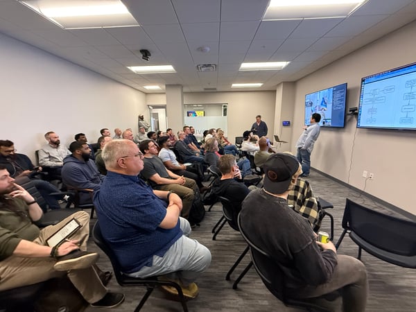 A group of people sitting in a conference room attending a technical presentation. A presenter stands at the front next to two large digital displays showing flowcharts and bullet points.
