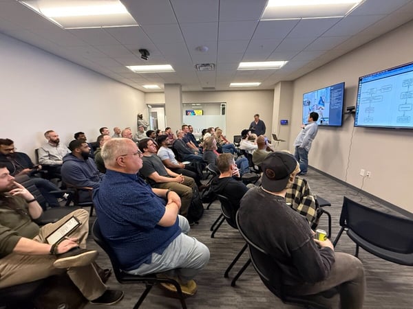 A group of people are seated in a conference room, attentively watching a presentation delivered by a man standing at the front next to two large screens.