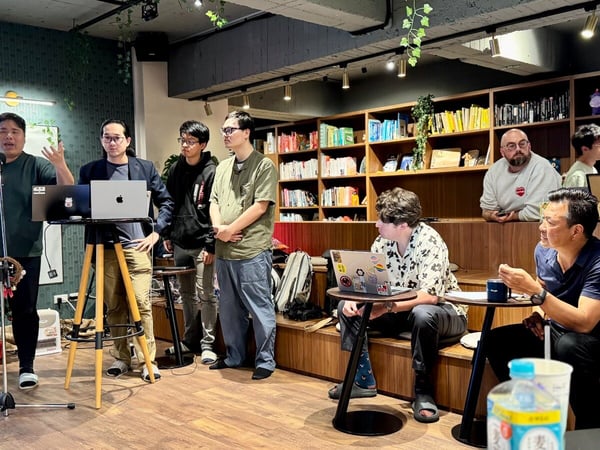 A group of nine men is gathered in a modern, cozy indoor space, decorated with a Christmas tree, where one man is giving a presentation using a microphone and laptops. The setting includes tiered wooden seating, bookshelves filled with books, and potted greenery.