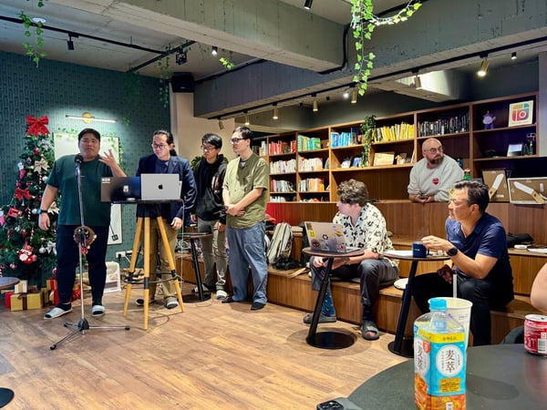 A candid photograph captures an indoor group gathering, likely a technical presentation or meeting, featuring a man speaking into a microphone while others stand nearby and several observe from tiered seating in a room decorated with bookshelves and Christmas elements.