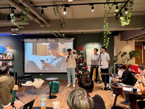 An indoor photograph capturing a presentation or informal meeting in a warmly decorated space, where a speaker addresses an audience in front of a large projection screen displaying a close-up image of a man using a smartphone.
