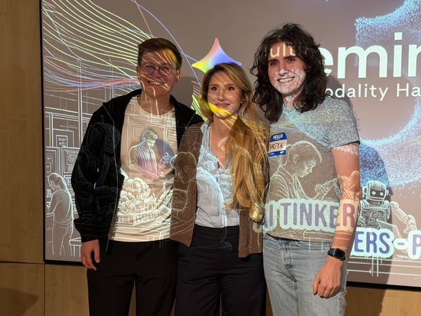 Three people stand together in front of a large screen where graphics and text related to a Gemini hackathon are projected onto both the screen and the people themselves.