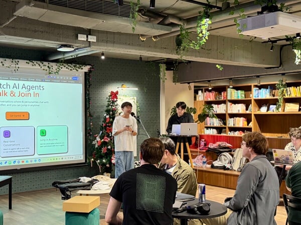 A photograph captures an indoor tech presentation or meetup where a speaker, holding a microphone, stands next to a large projected screen displaying information about 'Watch AI Agents Talk & Join In.' Several attendees are seated in the foreground and mid-ground, watching the presentation in a casual setting decorated with plants and bookshelves.