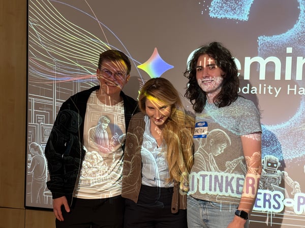 Three individuals posing for a photo in front of a projection screen displaying 'Gemini' and 'modality Hackathon' graphics, with some of the light patterns falling on their clothing.