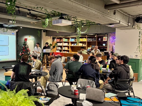 A group of people is attending an indoor presentation or meeting in a casual, co-working or lounge space, where two individuals are speaking near a large projection screen displaying four categorized content boxes. The room features exposed concrete ceilings, bookshelves, abundant greenery, and holiday decorations like a small Christmas tree.
