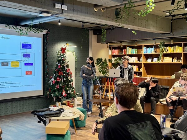 A photograph capturing a technical presentation or meetup taking place indoors, featuring a female presenter speaking into a microphone next to a screen displaying a 'System Architecture Diagram,' while several attendees watch in a cozy, decorated space.