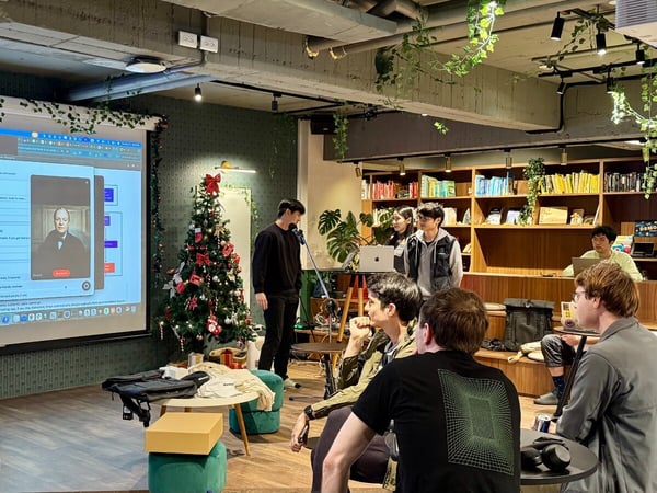 A photograph capturing an indoor presentation or meeting in a modern, plant-filled event space decorated with a Christmas tree, where a speaker addresses a seated audience while an application interface is displayed on a large projection screen.