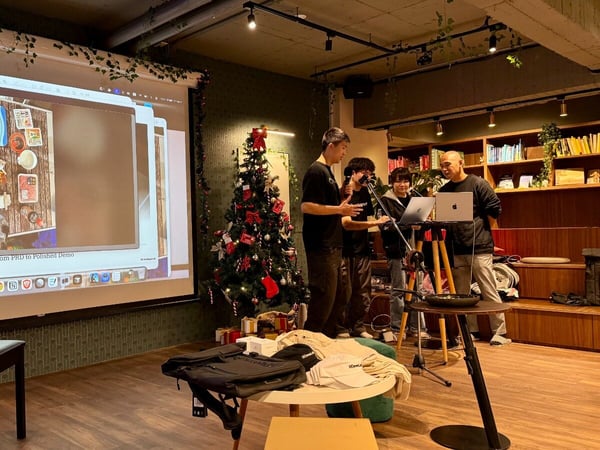This is a photograph of an indoor presentation or demo session, likely during the holiday season, where four individuals are gathered near a projection screen displaying images of food ingredients and the title "From PRD to Polished Demo." The setting features warm lighting, a decorated Christmas tree, and bookshelves.