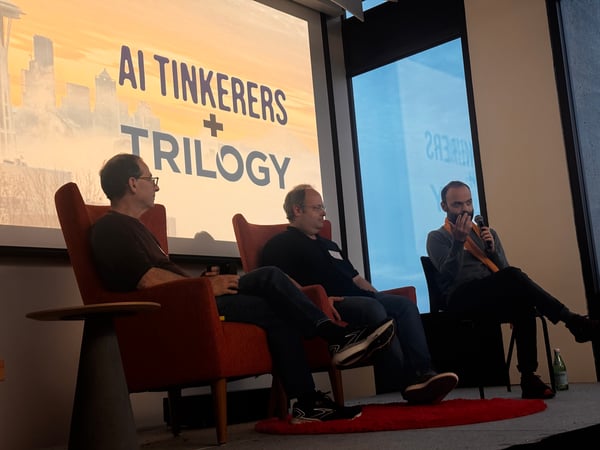 A photograph capturing a panel discussion featuring three men seated on a low stage, with a large projection screen behind them displaying the title 'AI TINKERERS + TRILOGY' over a faint cityscape silhouette.