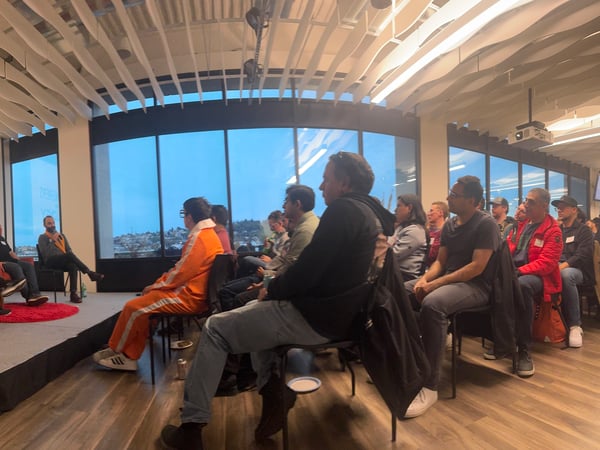 A wide-angle photograph captures an indoor panel discussion event where three speakers are seated on a stage in front of a large screen displaying the title 'AI TINKERERS + TRILOGY' to a seated audience.
