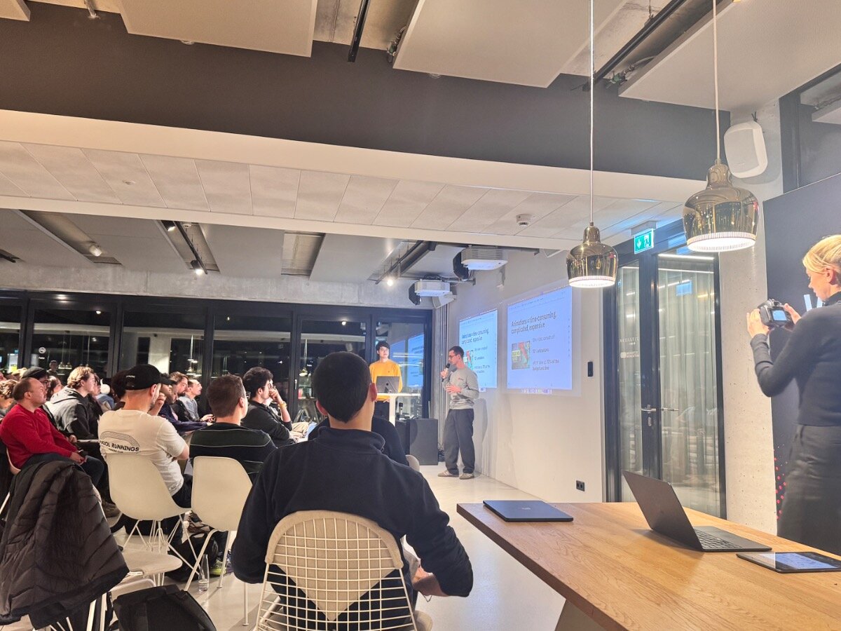 A candid photograph capturing an indoor presentation or technical meetup, showing two speakers standing in front of a projection screen while addressing a large audience seated in rows. A person on the right side is visible taking a photo of the scene.