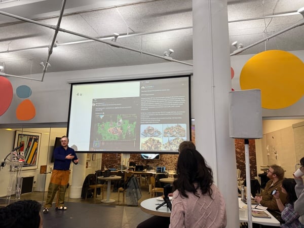 A man is giving a presentation in a modern office space, using a large projector screen that displays social media posts and game-related graphics.