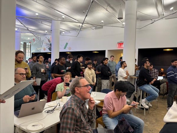 A group of people gathered in a modern, open-plan office or event space, many of whom are looking toward an off-camera presentation or demonstration.