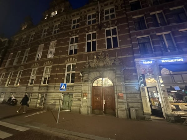 A nighttime street view featuring an ornate brick building with a large arched wooden entrance, located next to a brightly lit hotel and restaurant.