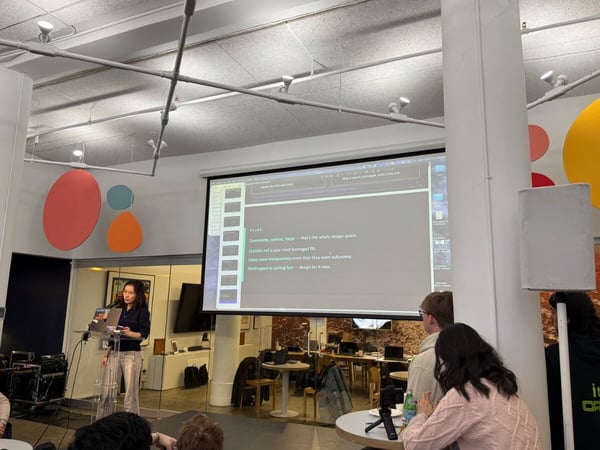 A woman stands at a podium giving a presentation in a modern office space with a large projector screen showing bullet points about design and AI.