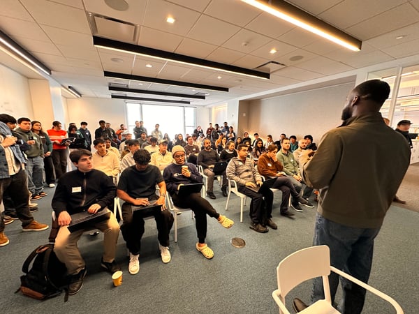 A man stands in the foreground with his back to the camera, addressing a large, diverse audience seated and standing in a modern office or classroom setting.